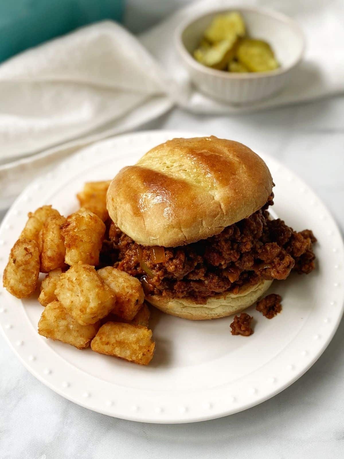 overhead shot of sandwich on a white plate with tater tots with a dish of pickles in the background