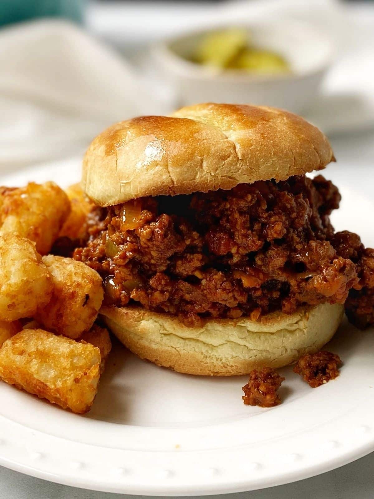 sandwich on a white plate with tater tots and a bowl of pickles in the background