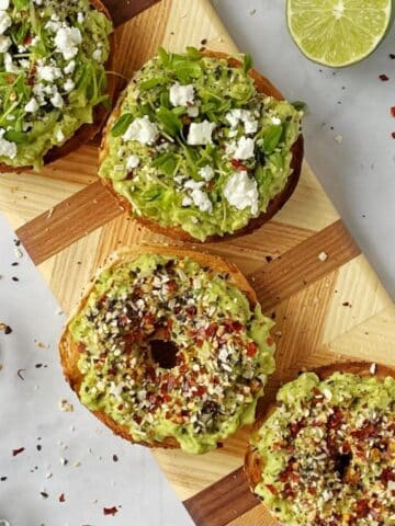 bagels with avocado and toppings on a cutting board.