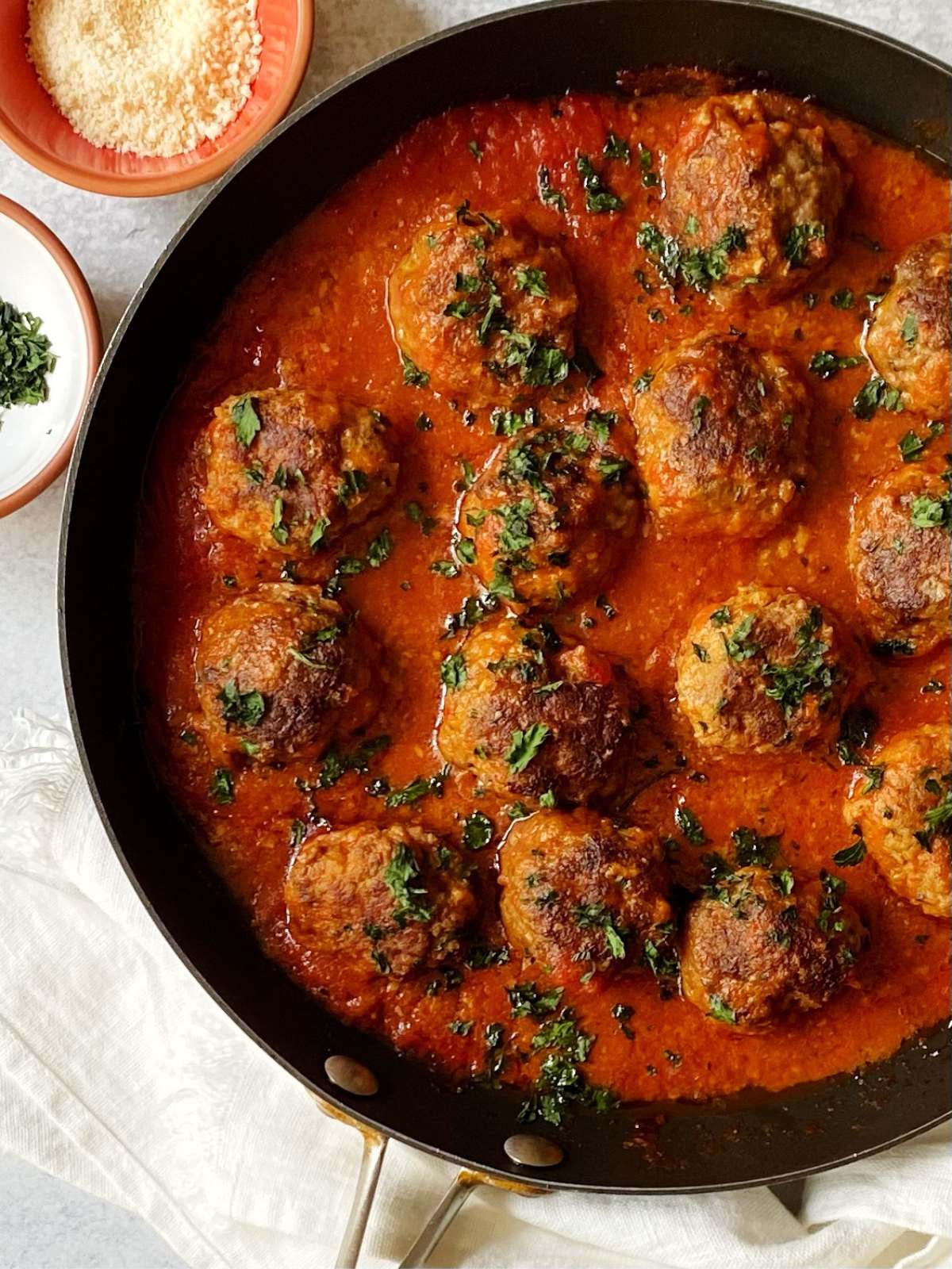 top-down shot of meatballs and sauce in a skillet next to small dishes of parsley and parmesan.