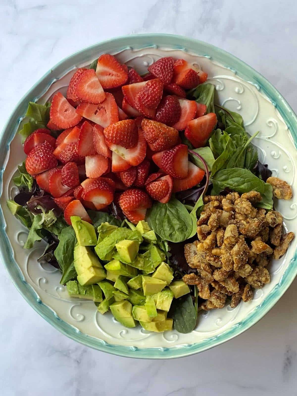 A bowl of lettuce topped with strawberries, avocado, and candied walnuts before tossing.
