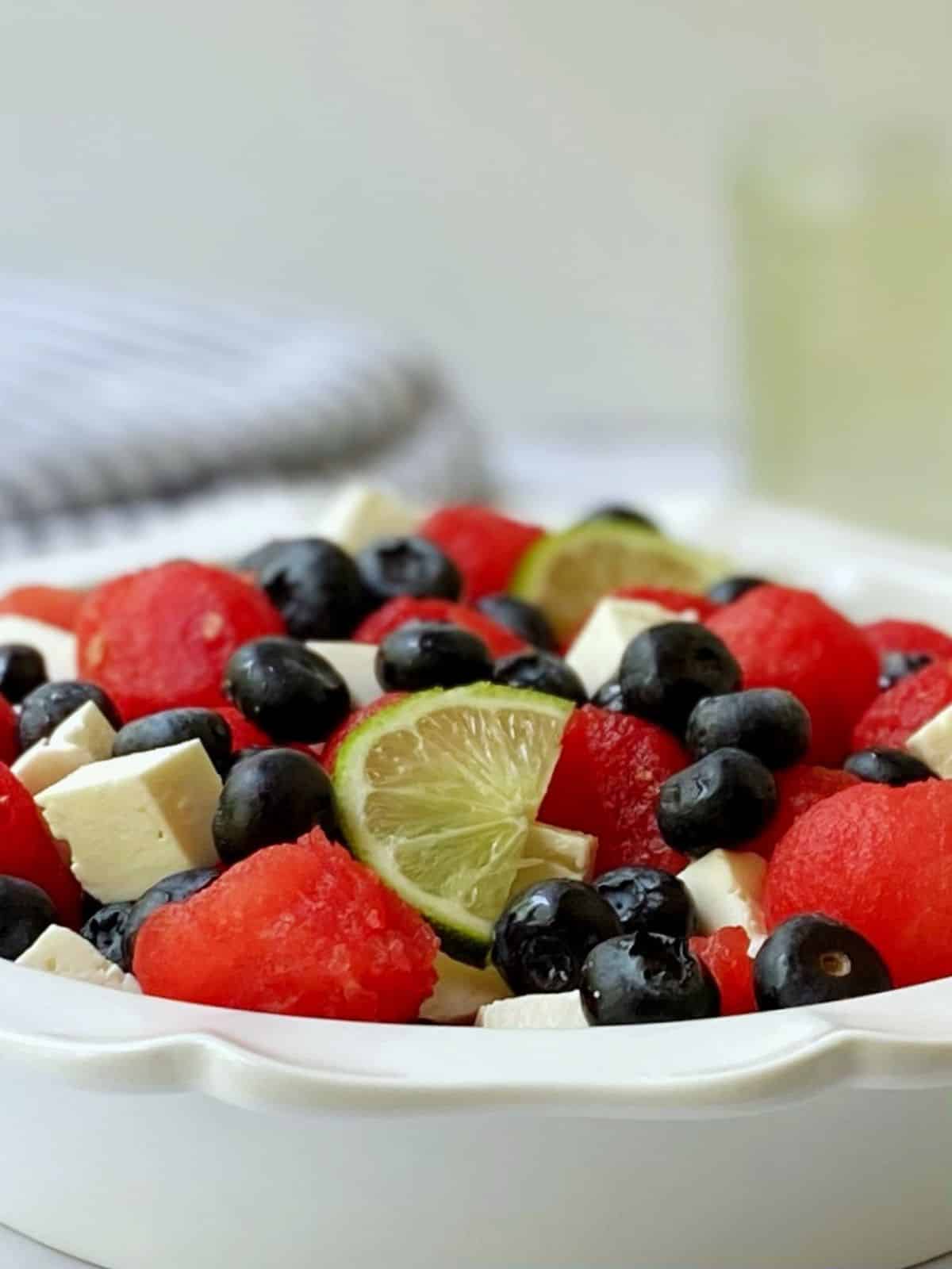 side view of watermelon feta salad in a white serving dish with a jar of extra simple syrup in the background.