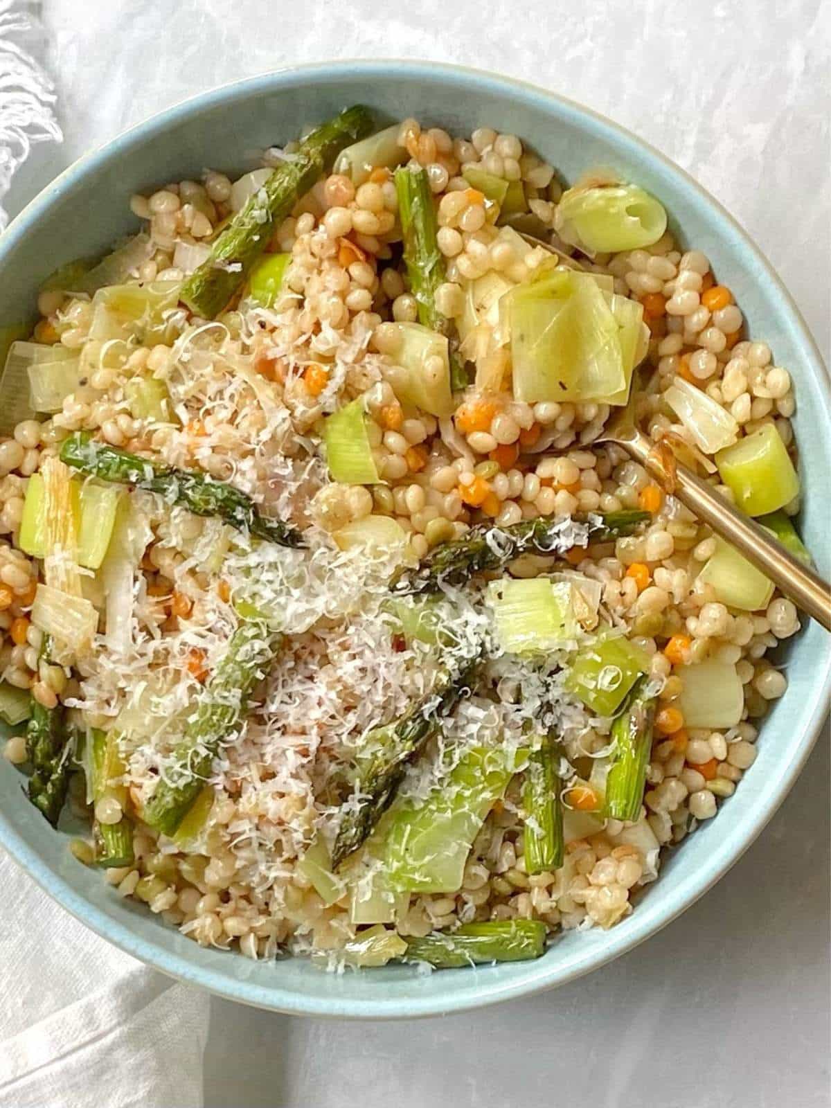 Gold serving spoon in a blue bowl filled with brown butter couscous, asparagus and leeks.