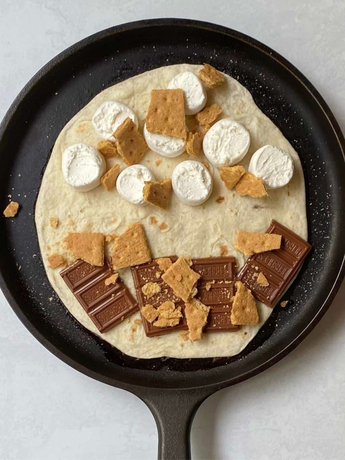 tortilla on a griddle topped with chocolate, marshmallows, and graham crackers.