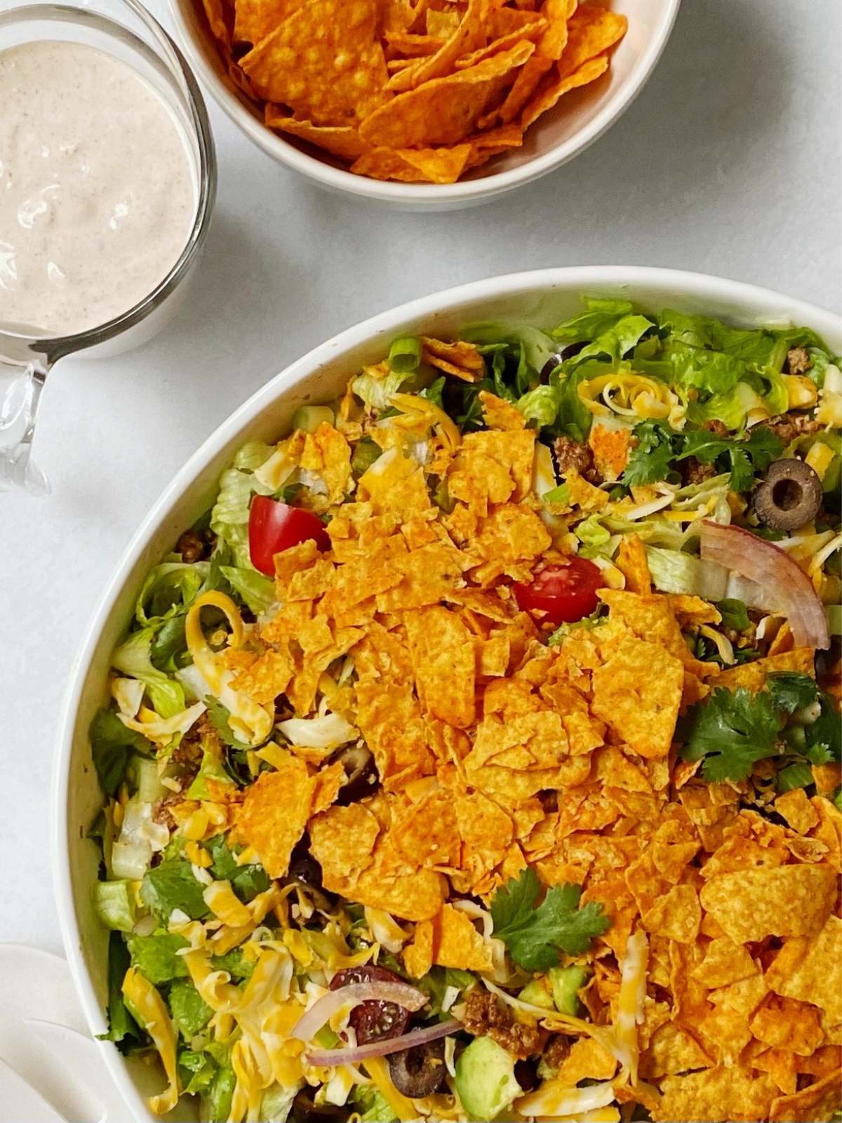 Large serving bowl of Doritos taco salad next to a pitcher of dressing and bowl of Doritos.