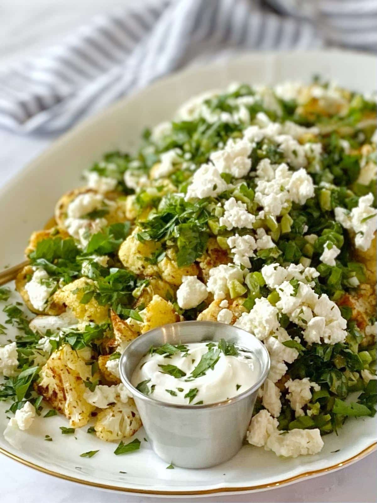 Close up of a platter of roasted cauliflower topped with olive, herbs, and feta.