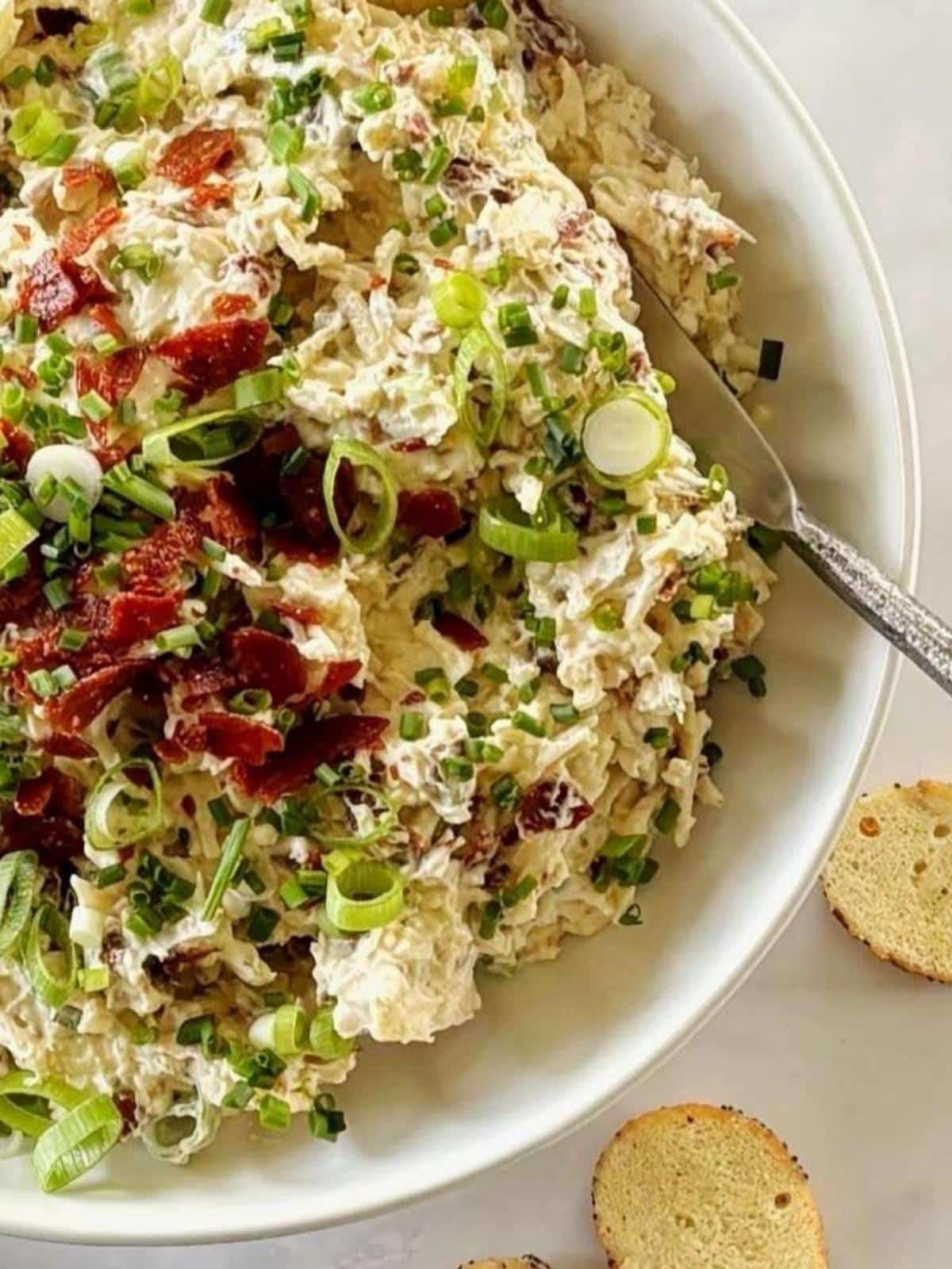 A spreader in a serving bowl of cold dip alongside bagel chips.