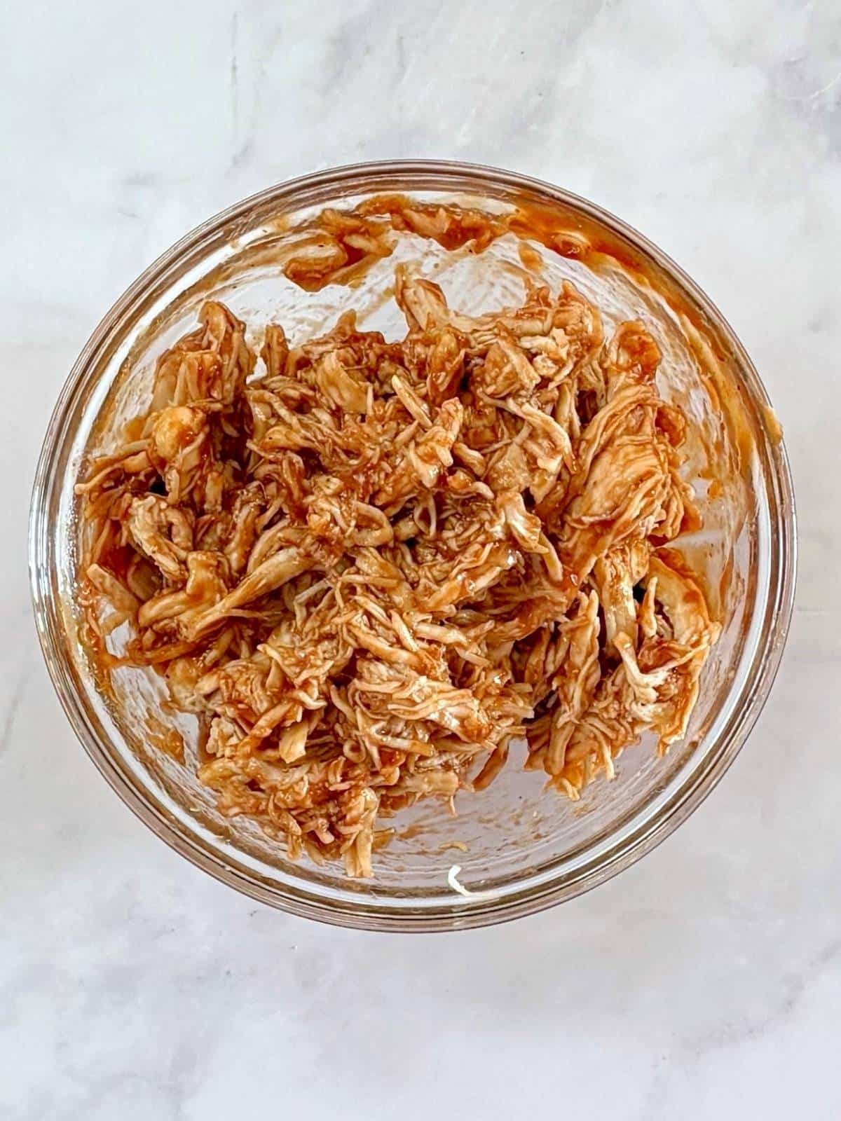 Shredded chicken tossed with BBQ sauce in a mixing bowl.