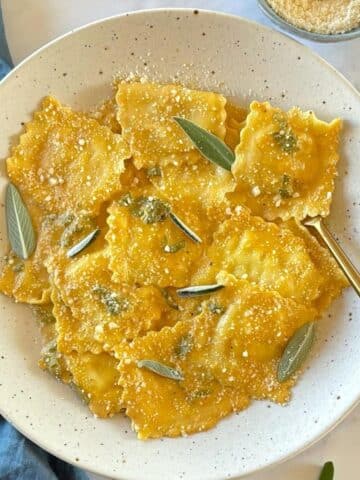A fork in a serving bowl of ravioli next to a small dish of Parmesan cheese.